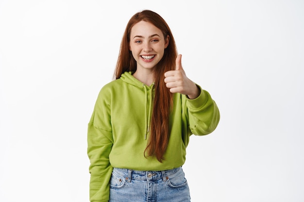 Very good. Smiling redhead teen shows thumbs up and looks satisfied, praise great deal, recommending store, make compliment, white background.