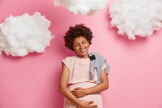 Очень счастливая улыбающаяся беременная афроамериканка нежно прикасается к животу и чувствует своего ребенка, покупает комбинезон для новорожденного, наслаждается моментом материнства и материнства, играет с драгоценным будущим будущим ребенком