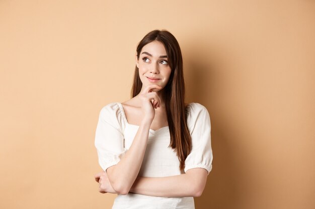 Very interesting. Pensive cute girl thinking and looking at upper left corner logo, smiling pleased, having good idea, standing on beige background