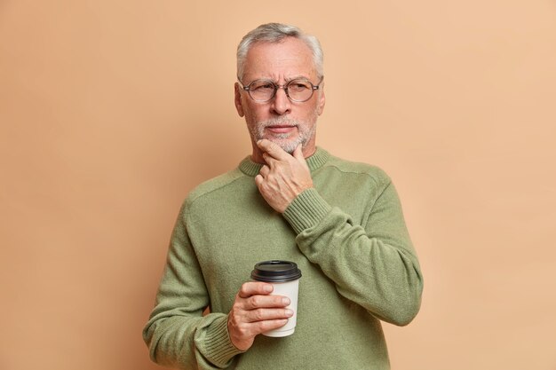 very serious mature European man holds chin looks pensively aside drinks coffee to go considers something important wears casual sweater and spectacles isolated over brown wall