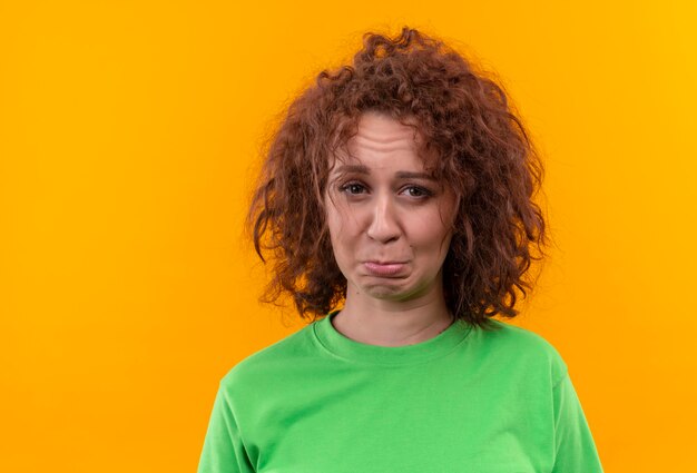 Very upset young woman with short curly hair in green t-shirt looking at camera frowning 