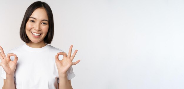 Very well excellent Smiling asian woman showing okay sign approval ok gesture looking satisfied recommending smth standing over white background