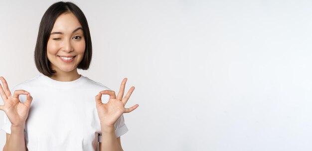 Very well excellent Smiling asian woman showing okay sign approval ok gesture looking satisfied recommending smth standing over white background