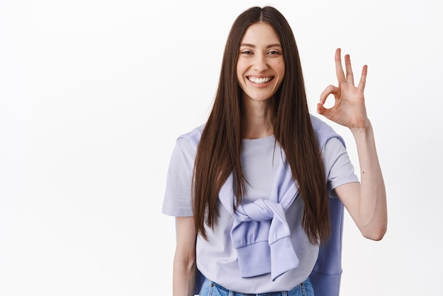 Very well good work Smiling brunette girl show OK okay sign in approval nod and agree give positive answer praise excellent choice standing in tshirt against white background