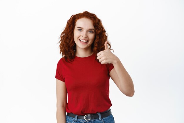 非常によく親指を上げて承認でうなずく笑顔を見せているポジティブな赤毛の女子学生の肖像画は、はいまたは申し分なく製品の承認をお勧めし、白い背景が好きです