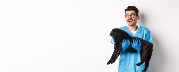 Free photo vet clinic concept happy male doctor veterinarian holding cute black pug dog smiling and looking lef