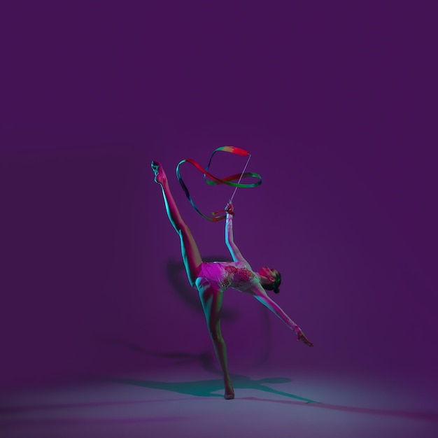 Яркий. молодая спортсменка, артистка художественной гимнастики танцует, тренируется с лентой, изолированной на фиолетовом студийном фоне в неоновом свете. красивая девушка практикуется с оборудованием. грация в исполнении Premium Фотографии