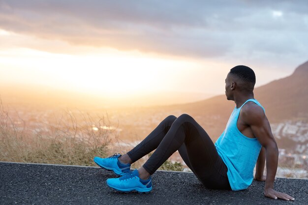 View of active male rests afer sprinting, sits at asphalt and leans on hands, dressed in activewear, blue sneakers, keeps gaze aside on panoramic picture of mountain nature, being full of energy