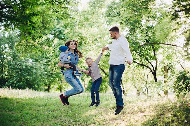Free photo view of active and positive family having fun and jumping in park together with kids mother and father embracing their two son and holding by hands concept of family relationship