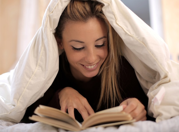 白い毛布の下で本を読んでいる美しい白人女性のビュー