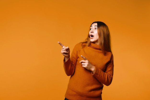 View of beautiful surprised redhead caucasian woman dressed in pullover, showing something with fingers on the left corner