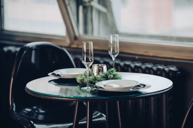 Вид на красиво оформленный круглый стол с натуральной еловой веткой, свечой, двумя флейтами, тарелками на фоне классического дивана в современной квартире.