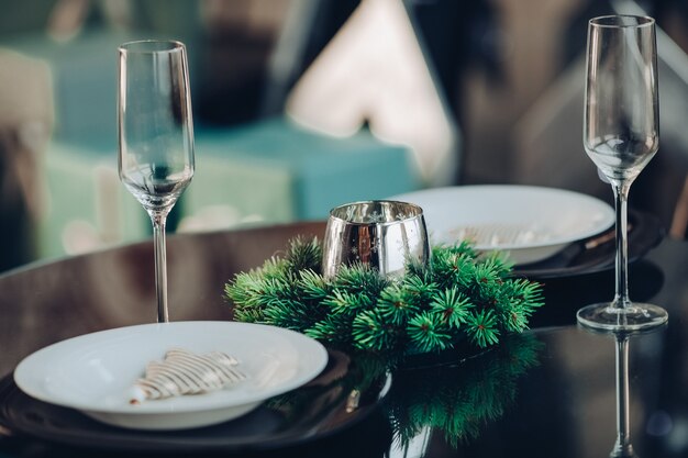 View over beautifully decorated round table with natural fir branch, candle, two flutes, plates against classic sofa in modern apartment.