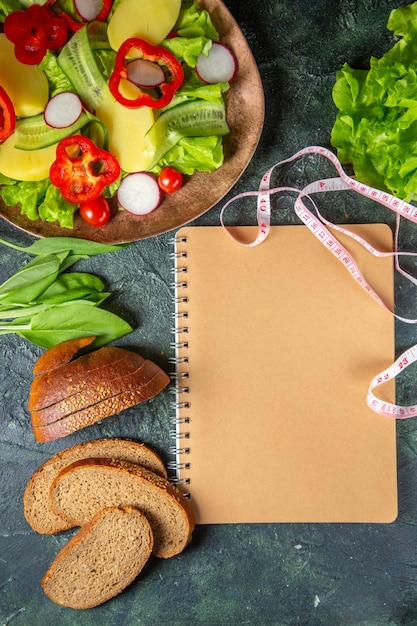 Above view of black bread slices fresh chopped vegetables on a plate and meters green bundle spiral notebook on dark colors surface