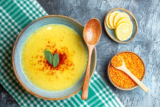 Above view a blue pot with tasty soup served with mint and pepper next to chopped lemon wooden spoon and yellow pea on blue background