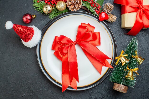 Above view of bow shaped red ribbon on dinner plate christmas tree fir branches conifer cone gift box santa claus hat on black background