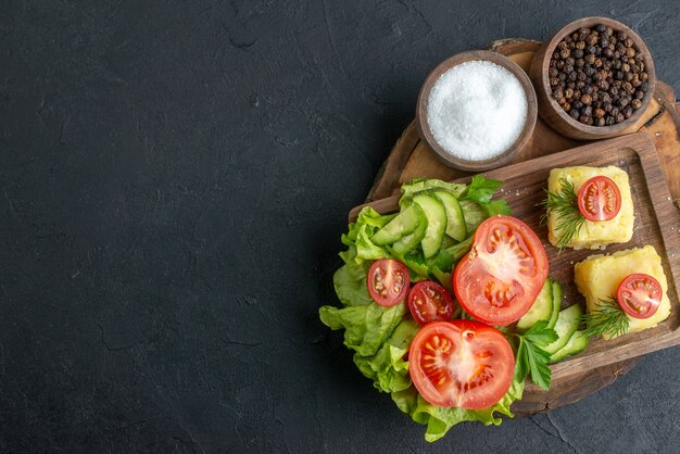 Above view of chopped and whole fresh vegetables cheese on cutting board and spices on the left side on black surface