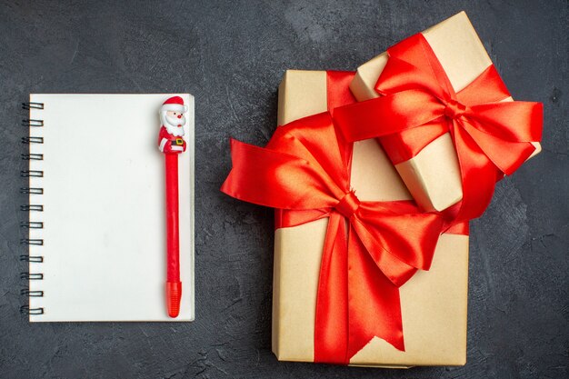 Выше вид на рождественский фон с красивыми подарками с бантовой лентой и блокнотом с ручкой на темном фоне