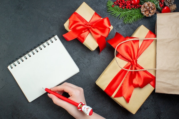 Above view of Christmas mood with beautiful gifts and fir branches conifer cone next to notebook with pen on dark background