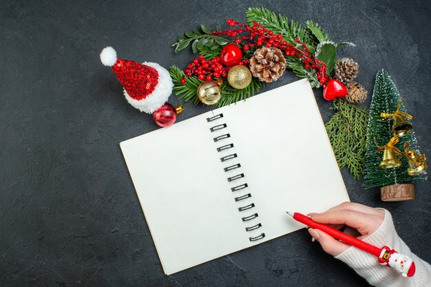 Above view of christmas mood with fir branches xsmas tree santa claus hat hand holding a pen on spiral notebook on dark background