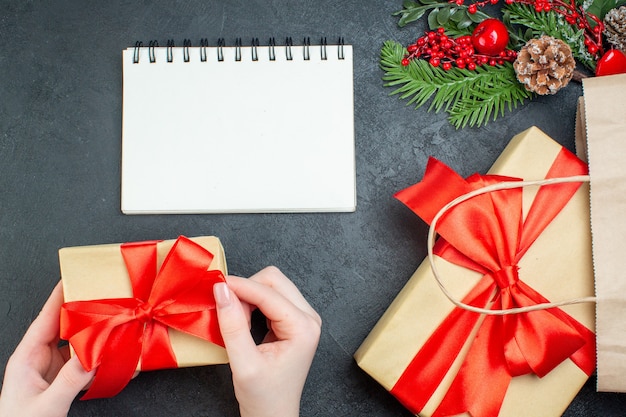 Above view of Christmas mood with hand holding one of beautiful gifts and fir branches conifer cone next to notebook on dark background