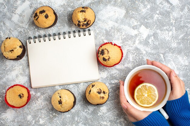 チョコレートと氷の表面にレモンと紅茶のカップを持っている手でおいしい小さなカップケーキの中で閉じたノートブックのビューの上