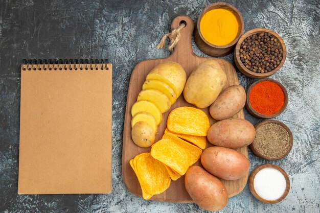 Above view of crispy chips and uncooked potatoes on wooden cutting board different spices and notebook on gray table