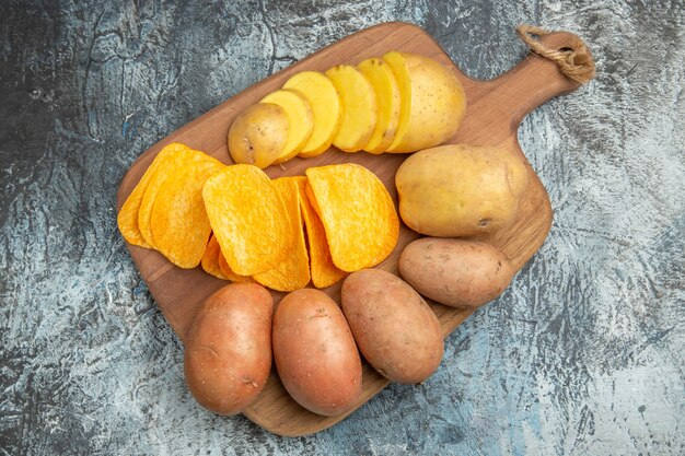 Above view of crispy chips and uncooked potatoes on wooden cutting board on gray table