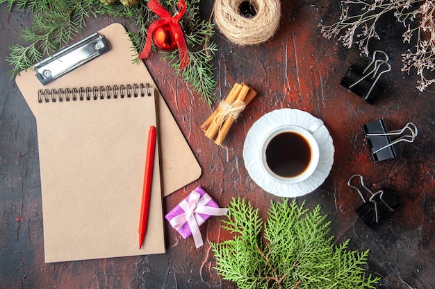 Free photo above view of a cup of black tea fir branches cinnamon limes conifer cones gift and notebook on dark background