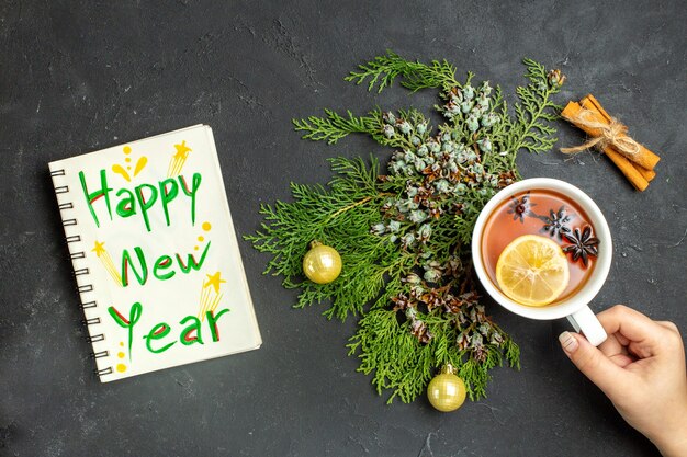 Above view of a cup of black tea xsmas accessories and cinnamon limes and notebook with happy new year inscription on black background