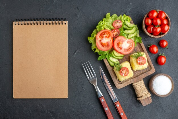 Above view of cut fresh tomatoes and cucumbers cheese on wooden board cutlery set salt notebook on black surface