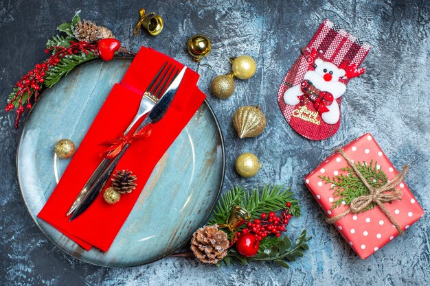 Above view of cutlery set with red ribbon on a decorative napkin on a blue plate and christmas accessories next to gift box and christmas sock on dark background