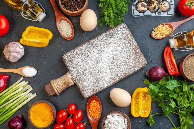 Above view of cutting board with flour among fresh vegetables different spices green bundles fallen oil bottle bread slices eggs on dark