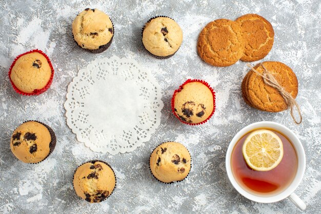 Above view of decorated napkin among delicious small cupcakes with chocolate and a cup of black tea with lemon stacked cookies on ice surface