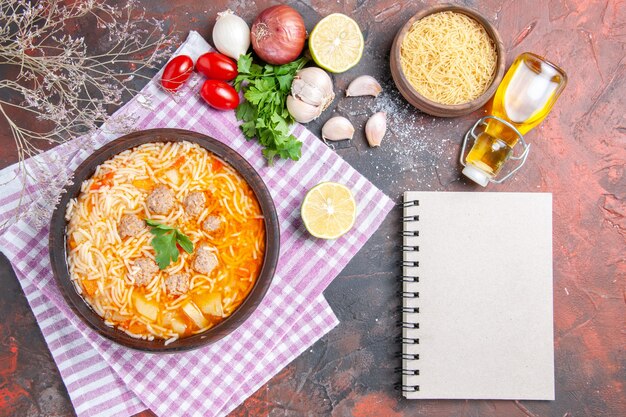 Above view of delicious chicken soup with noodles greens and spoon on pink stripped towel oil bottle garlic tomatoes lemon and notebook on dark background