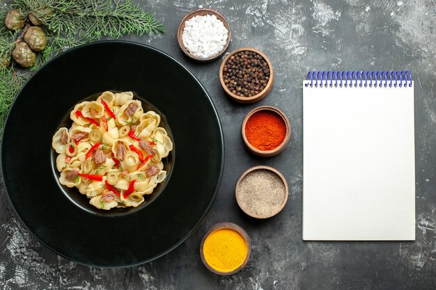 회색 배경에 있는 노트북 옆에 접시와 칼, 다른 향신료를 넣은 야채와 채소를 곁들인 맛있는 콘치글리의 전망