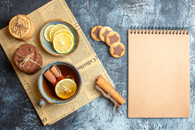 Above view of delicious cookies and a cup of black tea with cinnamon on an old newspaper next to spiral notebook on dark background