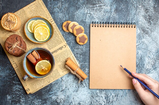 Above view of delicious cookies and a cup of black tea with cinnamon on an old newspapnd holding pen on spiral notebook on dark background