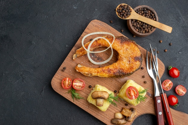 Above view of delicious fried fish and mushrooms tomatoes greens on wooden cutting board cutlery set pepper on black surface