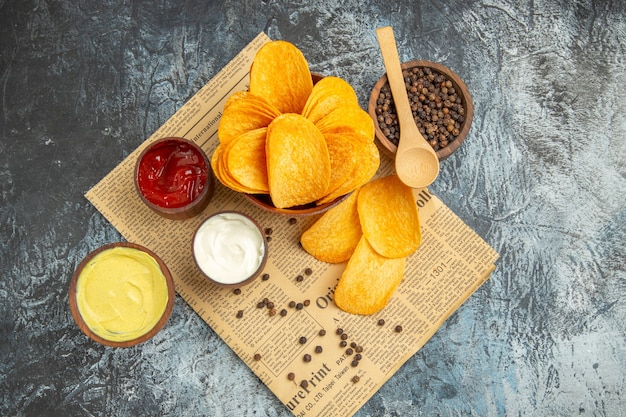 Above view of delicious homemade chips and pepper bowl mayonnaise ketchup and sauce with spoon on newspaper on gray table