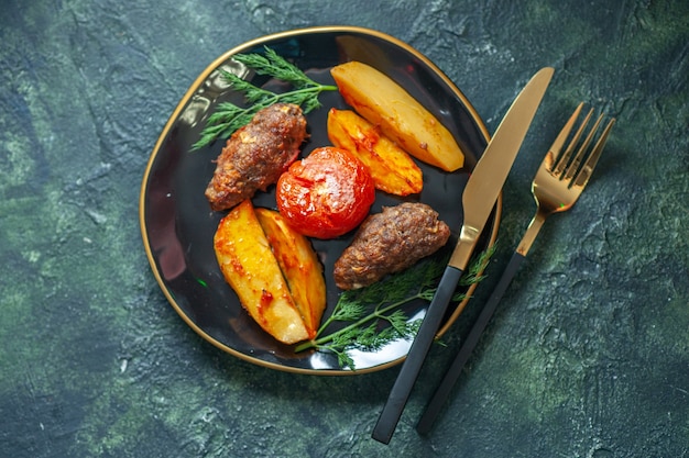 Above view of delicious meat cutlets baked with potatoes and tomatoes on a black plate served with green cutlery set on green black mix colors background