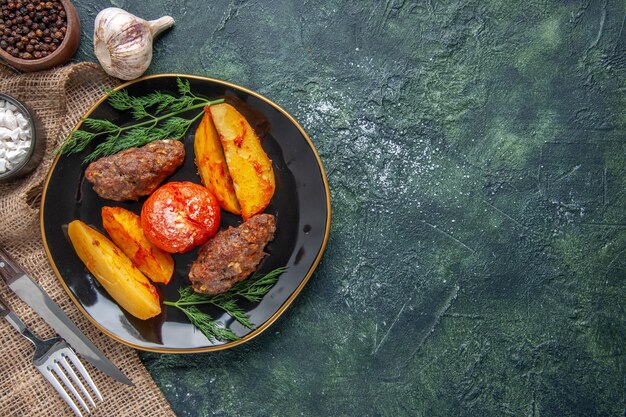 검정 접시에 감자와 토마토로 구운 맛있는 고기 커틀릿의 보기 위에 녹색 검정 혼합 색상 배경의 오른쪽에 있는 향신료 마늘 칼 붙이 세트