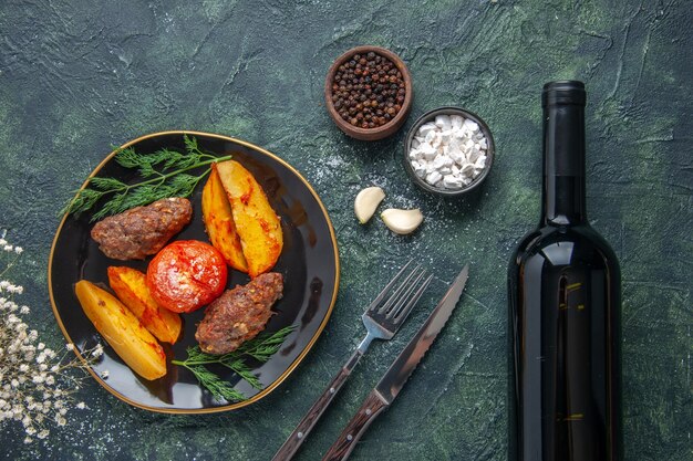 검은 접시에 감자와 토마토로 구운 맛있는 고기 커틀릿의 보기 위에 녹색 검정 혼합 색상 배경에 향신료 마늘 와인 병