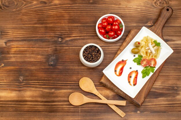 Above view of delicious pasta meal served with vegetables on a white plate on cutting board pepper tomatoes spoons on brown wooden background