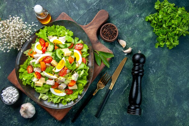 Above view of delicious salad with fresh ingredients on wooden cutting board spices oil bottle garlics cutlery set on black mix colors table