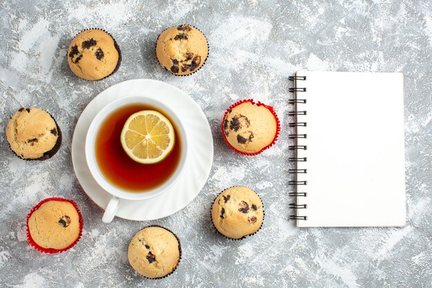Above view of delicious small cupcakes with chocolate around a cup of black tea next to notebook on ice surface