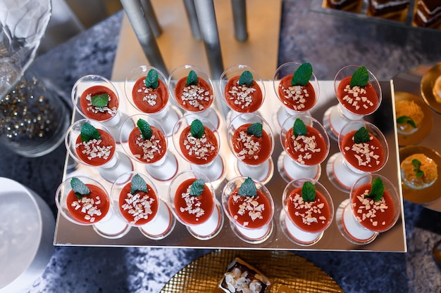 Above view of dessert sets in glasses cups which made with white and red jelly decorated by pieces of white chocolate and leaves of mint served on mirror plate on wedding candy table