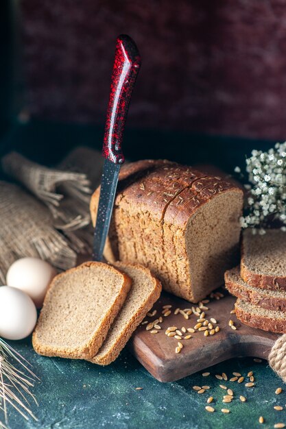 Above view of dietary black bread wheats on wooden cutting board knife flower eggs flour in bowl brown towel on mixed colors background