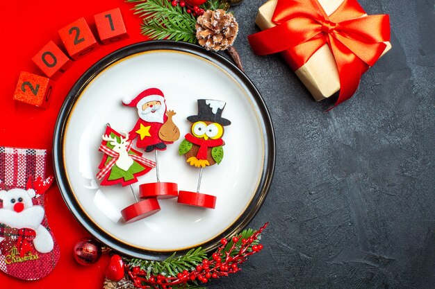 Above view of dinner plate decoration accessories fir branches and numbers christmas sock on a red napkin next to gift on a black table