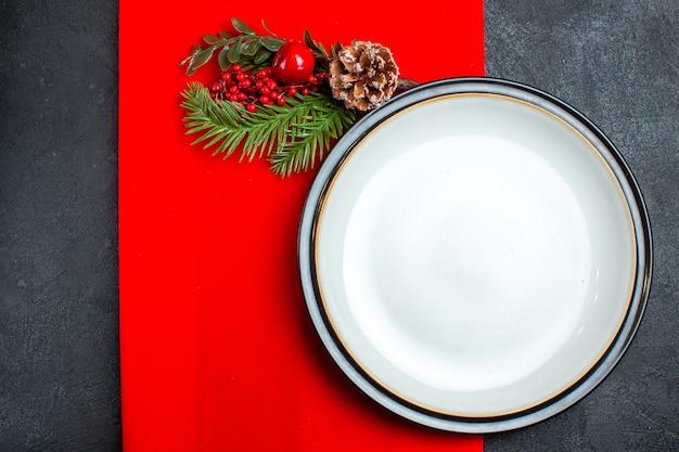 Above view of empty plates and fir branches with decoration accessory conifer cone on a red napkin on a dark background